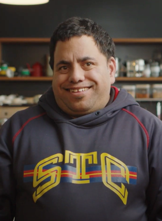 Young Maori man facing camera with short dark brown hair, brown eyes and smiling at camera. He has a dark blue sweatshirt with STC in yellow on the front. there are shelves with jars behind him.