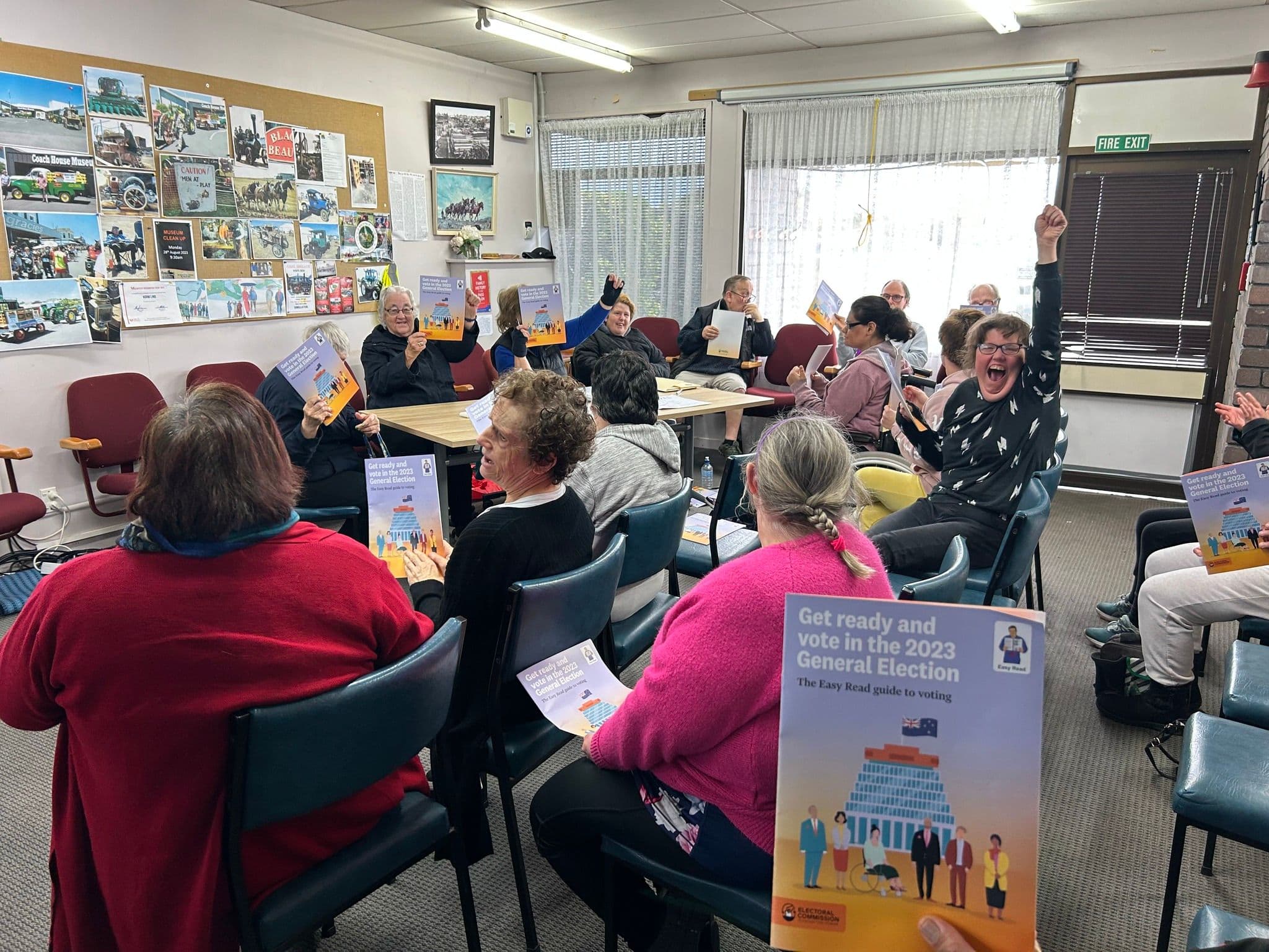 people sitting in a room holding an easy read document for "Get ready and vote in the 2023 General Election" One lady has her hand up excited.
